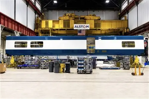 Гладкая металлическая обшивка новых вагонов-кафе Amtrak в белую и темно-синюю полоску стоит на производственной линии завода Alstom в окружении ярко-желтого и красного стального промышленного оборудования, серых и желтых пластиковых контейнеров и другого производственного оборудования.