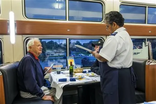 Официантка в белой рубашке с фирменными погонами Amtrak и синем фартуке принимает заказ от старшего джентльмена с белыми волосами и синей толстовкой в традиционном вагоне-ресторане Amtrak, укомплектованном белыми скатертями, салфетками из синей ткани и зеленым вином. бутылка, плетеная корзинка для хлеба и вид на парковку вокзала в сумерках.