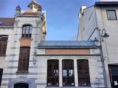 Каменное здание в стиле модерн с кирпичами разных оттенков серого и оранжевой вывеской с надписью Jardin d’Enfants-Kindertuin; есть серая крыша, освинцованные окна и трубы, и он примыкает к таким же зданиям.