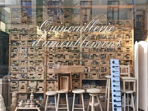 Витрина магазина с надписью Quincaillerie d'ameublement белым курсивом; в окне ряды и ряды деревянных полок, украшенных ручками, ручками и молоточками; также представлены табуретки и другие деревянные предметы.