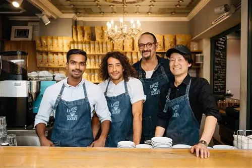 Четверо мужчин в джинсовых фартуках с вышитым логотипом kaffeerosterei стоят за деревянной стойкой и улыбаются в камеру. Позади них, не в фокусе, полки с пакетами кофейных зерен и люстра, свисающая с богато украшенного потолка.
