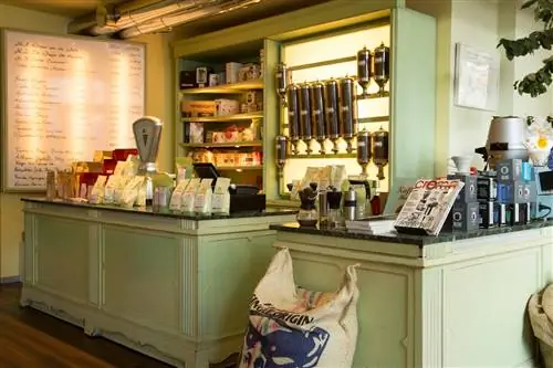 Интерьер Vits der Kaffee, жаровни и кафе. Стены и прилавок с мраморной столешницей окрашены в бледно-зеленый цвет. Прилавок и полки за ним заставлены кофейными продуктами, а слева есть доска, на которой показаны различные доступные смеси кофе.