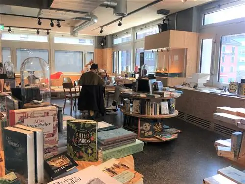 Круглые столы-витрины заставлены книгами для продажи в Космосе: за ними работают покупатели, а на стенах есть окна, выходящие на другие здания снаружи.