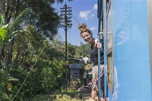 Женщина свисает из двери поезда, улыбаясь в камеру, расположенную из окна поезда. Поезд ярко-синего цвета, солнечный день в Элле, Шри-Ланка. Рядом с дорожками растут тропические деревья.