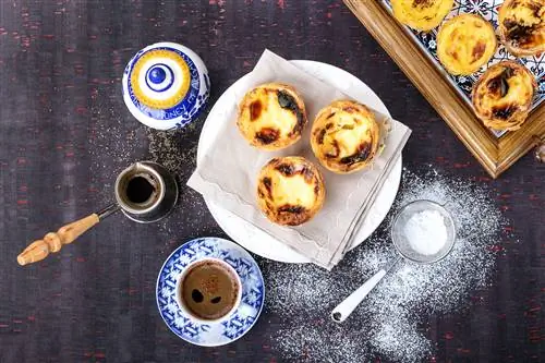 Традиционная португальская выпечка Pastel de Nata подается с кофе и медом на подносе над деревенской деревянной доской. Рядом с выпечкой на доске также есть сито с сахарной пудрой.