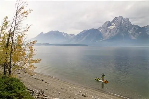 Мужчина гребет на байдарке по туманному озеру на фоне возвышающейся горы в национальном парке Гранд-Титон; Катание на байдарках стоя