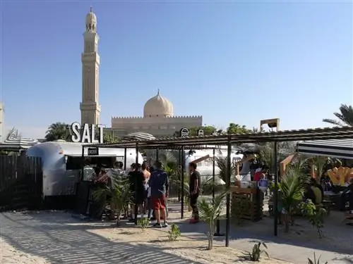 Уличный фуд-трак с солеными бургерами на пляже Кайт в Дубае. К небольшому металлическому трейлеру ведет крытая дорожка, где в солнечный день стоит очередь из нескольких человек. На заднем плане видна белая мечеть.