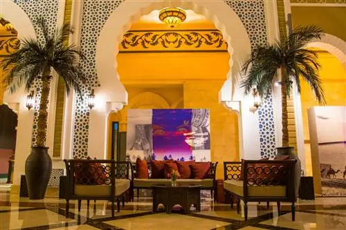 Вестибюль отеля в Дубае, Объединенные Арабские Эмираты. Деревянные диваны с темно-красными подушками расставлены под искусно выполненной аркой, через которую можно увидеть произведения современного искусства. Арка окружена высокими пальмами в горшках.