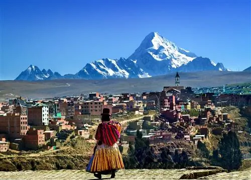 Женщина в традиционном боливийском платье идет по высокому хребту в Ла-Пасе, на фоне города и заснеженной горы. Боливия.
