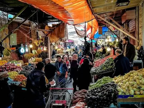 Фрукты и овощи сложены высокими кучами по обеим сторонам узкого прохода под крытым рынком; покупатели толкают тележки или несут сумки, а продавцы стоят позади своих товаров на приподнятых платформах; Бесплатные развлечения в Аммане, Иордания