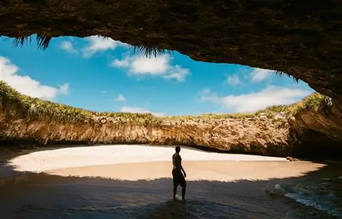 La Playa del Amor: мужчина стоит на пляже, который находится в круглой пещере и окружен скалой со всех сторон. Сверху виден кусок голубого неба с редкими пушистыми облаками. La Playa del Amor: мужчина стоит на пляже, который находится в круглой пещере и окружен скалой со всех сторон. Сверху виден кусок голубого неба с редкими пушистыми облаками.