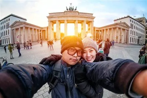 Пара делает селфи перед Бранденбургскими воротами в Берлине на закате. Мужчина протягивает руки, чтобы держать камеру, а женщина обнимает его. На обоих теплые пальто и шерстяные шапки.