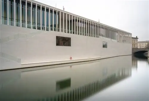 Фотография новейшего музея Музейного острова, Галереи Джеймса-Саймона, сделанная из-за воды. Здание очень современное, с большими кремово-бежевыми кирпичами, создающими плавные линии. Окна закрыты равномерно расположенными колоннами/.