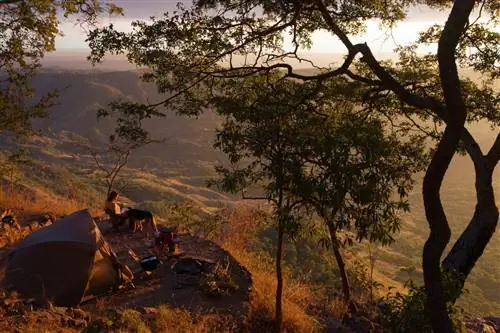 Женщина сидит в походном кресле рядом со своей собакой на плоском круглом участке кемпинга, который расположен на крутом склоне с видом на долину далеко внизу; свет золотой, скорее всего на закате