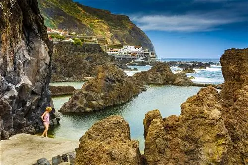 Женщина в розовом жилете и шортах стоит на краю естественного бассейна из вулканической породы рядом с океаном; дальше деревня и высокие скалы.