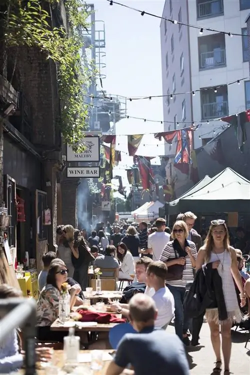 Посетители сидят на скамейках снаружи на одной стороне переулка, а торговые прилавки вдоль стен на другой стороне.