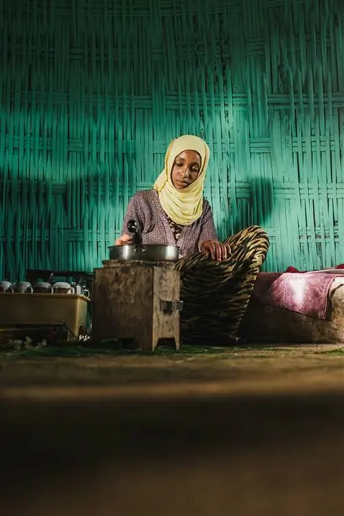 Эфиопская девушка готовит кофейную церемонию; на ней желтый платок и она сидит перед бирюзовой стеной.