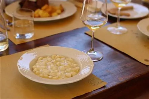 Тарелка сливочных ньокки подается на белой тарелке с другими блюдами, стаканами воды и белым вином, размытыми на заднем плане на темном деревянном столе