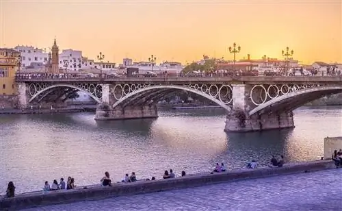 Закат над каменным мостом Триана и рекой Гвадалквивир в Севилье; на переднем плане люди сидят на каменной стене у реки.