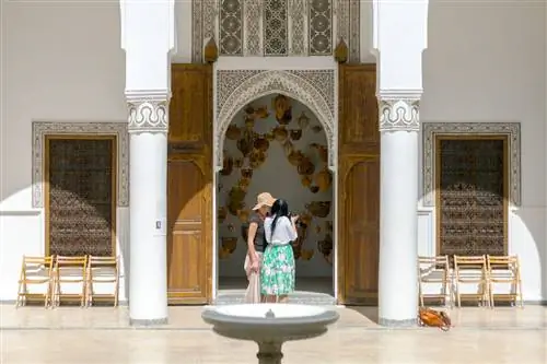 Двор Dar Bellarj, Марракеш, Марокко. На переднем плане небольшой каменный фонтан, за ним две женщины смотрят в камеру, обрамленную аркой высокой украшенной двери. Через дверь видно, что в воздухе висят глиняные чайники.
