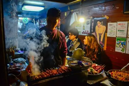 Японец жарит мясо для барбекю для клиентов, ожидающих на заднем плане в небольшом продуктовом ларьке в Омойдэ Йокочо.