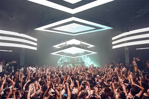 Большая толпа людей танцует под DJ Eric Prydz в мегаклубе Hï Ibiza. На потолке большие белые квадратные светильники, освещающие толпу внизу, которая в основном держит руки в воздухе и спиной к камере. На сцене зеленые и синие огни освещают ди-джея, который носит черную кепку перед деками.