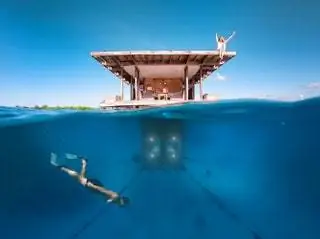 Мужчина ныряет перед окнами подводной комнаты Manta Resort, а женщина машет рукой с крыши.