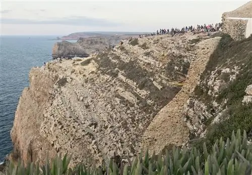 Десятки людей смотрят, как солнце исчезает за горизонтом на скале рядом с маяком Кабо-де-Сан-Висенте.