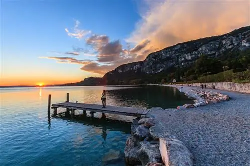 Вид на один из берегов Лаго-ди-Гарда на закате с людьми на песчаном пляже и на деревянном пирсе.